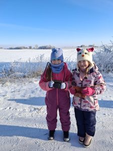 Dwoje dzieci w zimowych kombinezonach stoi na zaśnieżonym terenie i trzyma małe aparaty fotograficzne; w tle widoczne pole i krzewy pokryte szronem.