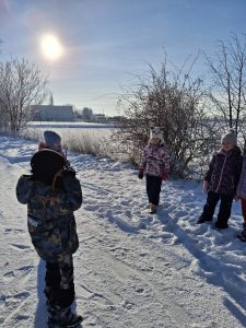 Dziecko fotografuje grupę dzieci stojących na zaśnieżonym terenie; w tle krzewy pokryte szronem, budynek i nisko świecące słońce.