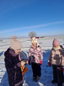 Troje dzieci na ośnieżonej przestrzeni ogląda aparat fotograficzny; w tle rozległe, zimowe pole i pojedyncze drzewo.