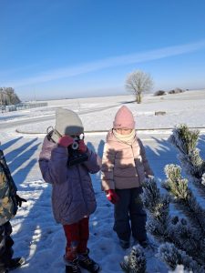 Dwoje dzieci w zimowych kurtkach stoi przy ośnieżonym krzewie; jedno dziecko fotografuje krzew aparatem.