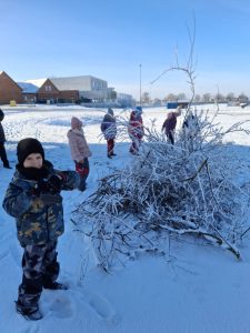 Kilka dzieci w zimowych ubraniach znajduje się na zaśnieżonym terenie obok sterty ośnieżonych gałęzi; jedno dziecko fotografuje scenę.