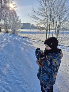 Dziecko w zimowej kurtce i czapce stoi na ośnieżonej drodze i trzyma aparat fotograficzny; w tle drzewa i budynek.