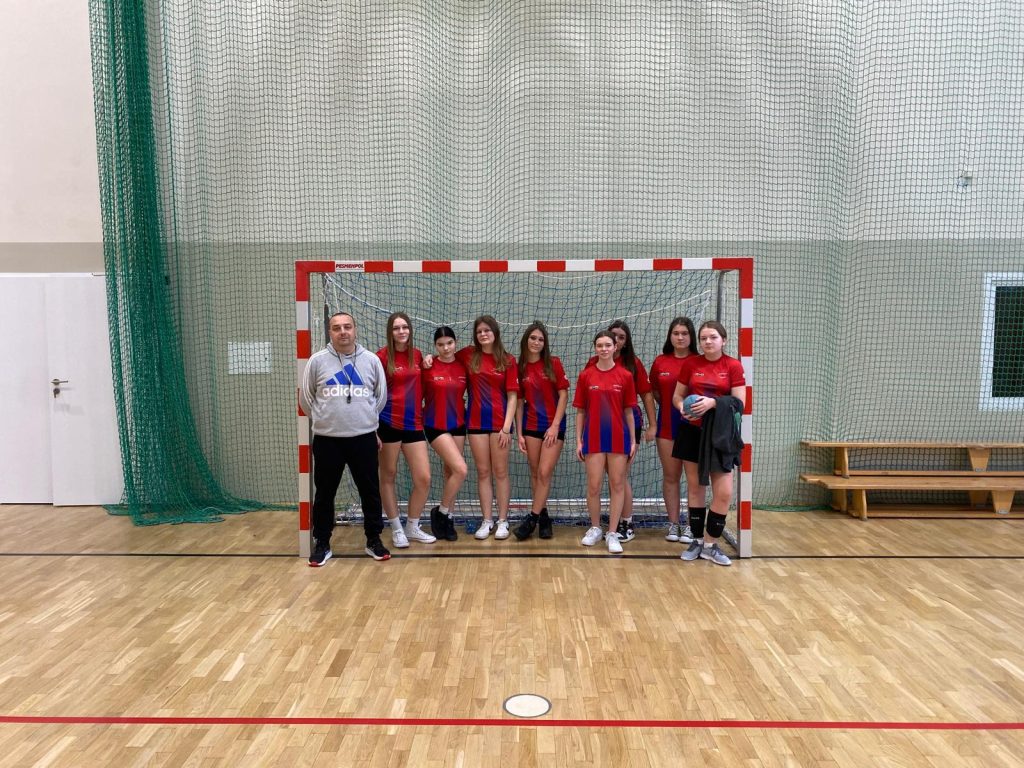 Grupowe zdjęcie drużyny piłki ręcznej w hali sportowej. Przed bramką stoi dziewięć zawodniczek w czerwono-niebieskich koszulkach sportowych oraz trener w jasnej bluzie. Zawodniczki mają stroje sportowe i obuwie halowe; jedna z nich trzyma piłkę. W tle widoczna jest siatka bramki, zielona siatka ochronna oraz ławki przy ścianie. Zdjęcie wykonane na drewnianej podłodze boiska.