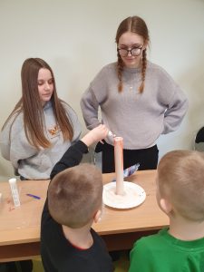 Uczeń oddziału przedszkolnego wlewa do wysokiej tuby z pianą ciecz z małej butelki. Po drugiej stronie stołu stoją dwie uczennice klasy 8 przyglądające się jego poczynaniom.