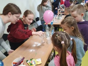 Uczniowie oddziału przedszkolnego oglądają eksperyment przygotowany przez dwie uczennice klasy 8. Na stoliku między nimi widoczny jest balon naciągnięty na plastikową butelkę.