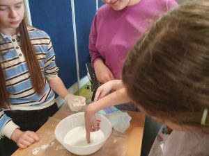 Uczennica z oddziału przedszkolnego wkłada palec do miski z cieczą nienewtonoską. Obok stoją uczennice klasy ósmej uwaznie ją obserwując.