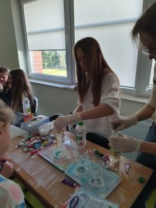 Uczennice klasy 7 wykonują eksperyment podczas którego w kolbie mieszają się ciecze o różnych kolorach.