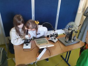 Dwie uczennice siedzą przy stoliku z mikroskopami. Jedna uważnie przygląda się zamieszczonej pod nim próbce.