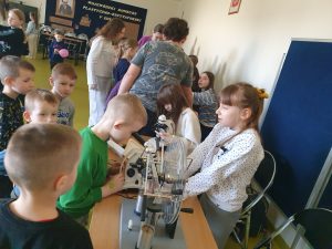 Dzieci z klas 0–3 oglądają preparaty przez mikroskopy, a starsze uczennice wyjaśniają działanie sprzętu.