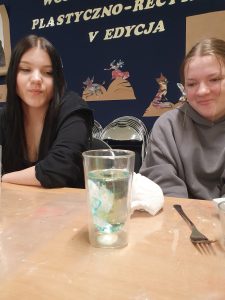 Dwie uczennice klasy 8 siedzą przy stoliku obserwując szklankę, w której mieszają się wlane do środka barwniki.