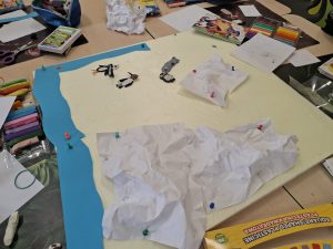 Etap pracy nad makietą: białe kartki imitujące lód przypięte pinezkami, figurki zwierząt i materiały plastyczne na stole.
