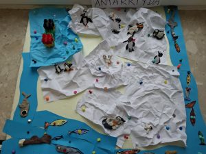 Makieta Antarktydy wykonana przez dzieci: białe kartki jako lód, niebieskie elementy jako ocean oraz figurki pingwinów i fok.