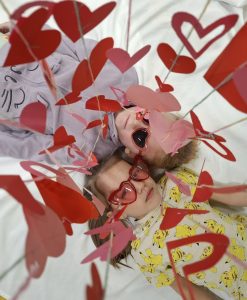 Dwoje dzieci leży na białym tle, patrząc w górę; mają czerwone okulary w kształcie serc, a nad nimi wiszą papierowe serca w odcieniach czerwieni i różu.