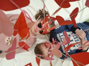 Dwoje dzieci leży na białym tle pod dekoracją z papierowych serc; mają czerwone okulary w kształcie serc.