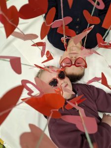 Dwoje dzieci leży obok siebie na białym tle, w czerwonych okularach w kształcie serc, otoczonych papierowymi sercami.