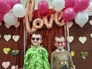 Dwoje dzieci stoi przed walentynkową dekoracją z balonów i napisem „love”; oboje mają czerwone okulary w kształcie serc.