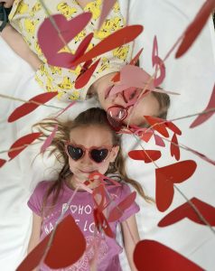 Dwie dziewczynki leżą na białym tle, patrząc w górę przez wiszące papierowe serca w kolorach czerwonym i różowym.