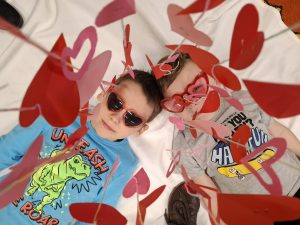 Dwoje dzieci leży na białym tle; mają czerwone okulary w kształcie serc, a nad nimi widoczne są wiszące papierowe serca.