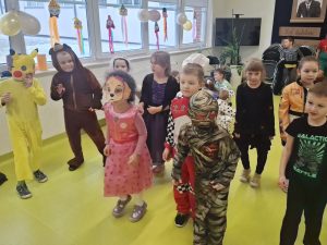 Dzieci z klas 0–III w kolorowych przebraniach karnawałowych tańczą na sali teatralnej udekorowanej balonami i girlandami.