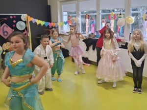Grupa dzieci w strojach bajkowych i baletowych bawi się podczas balu karnawałowego na sali teatralnej.