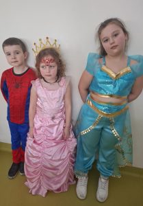 Trójka dzieci w strojach karnawałowych, w tym księżniczki i postaci z bajki, stoi przy ścianie sali teatralnej.