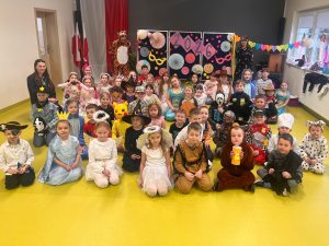 Duża grupa uczniów klas 0–III w różnorodnych przebraniach karnawałowych siedzi i klęczy do wspólnego zdjęcia na sali teatralnej wraz z wychowawczyniami.