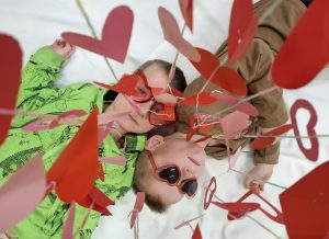 Dwoje dzieci leży na białym tle, mają czerwone okulary w kształcie serc; nad nimi znajdują się papierowe serca na sznurkach.