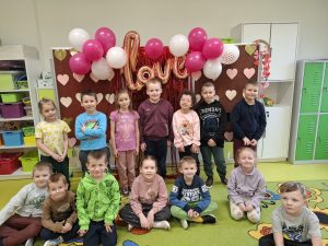 Grupa dzieci z oddziału przedszkolnego pozuje przed walentynkową dekoracją z balonów, czerwonych girland i napisu „love”.