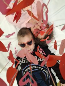 Dwoje dzieci leży na białym tle pod wiszącymi papierowymi sercami; jedno dziecko uśmiecha się, drugie patrzy w górę.