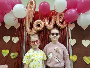 Dwie dziewczynki stoją przed tłem z napisem „love”, balonami i papierowymi sercami; mają czerwone okulary w kształcie serc.