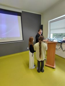 Przedstawicielka sanepidu przy mównicy rozmawia z uczennicami, pokazując treści na laptopie w trakcie zajęć profilaktycznych.