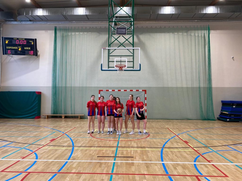 Drużyna dziewcząt w czerwono-niebieskich strojach sportowych stoi na środku sali gimnastycznej przed bramką i koszem do koszykówki.