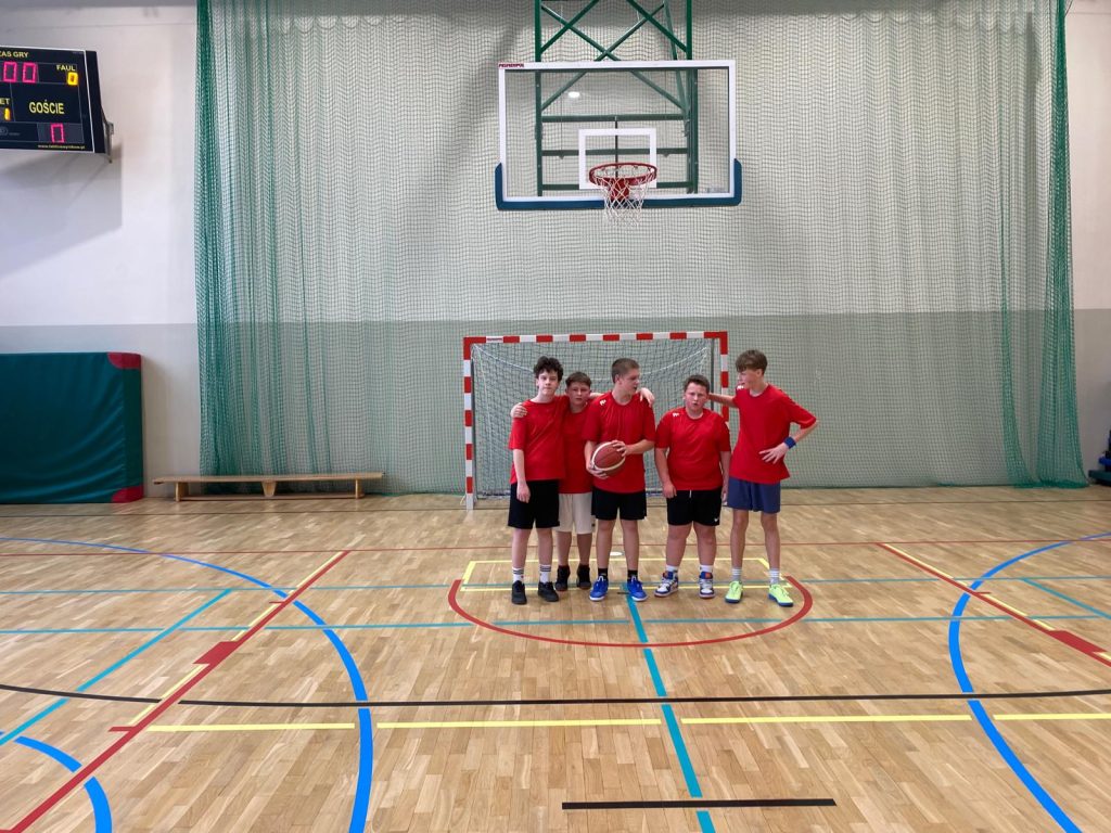 Drużyna chłopców w czerwonych koszulkach i czarnych spodenkach pozuje na środku sali gimnastycznej, jeden z zawodników trzyma piłkę do koszykówki.