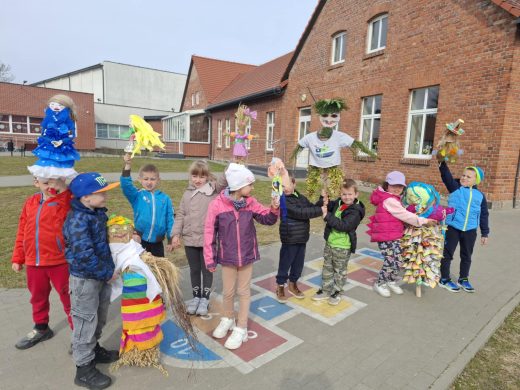 Dzieci z oddziału przedszkolnego stoją przed budynkiem szkoły i trzymają kolorowe kukły symbolizujące zimę, przygotowane na powitanie wiosny.