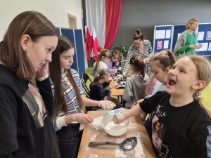 Uczennice klasy 8 pokazują doświadczenie z cieczą nienewtonowską przy stole. Jedna z młodszych uczennic wkłada rękę do miski z białą masą, reagując emocjonalnie. Na stole leżą rękawiczki, łyżka cedzakowa i miska z mieszaniną. W tle inne dzieci oglądają mikroskopy i stanowiska doświadczalne.