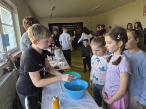 Uczeń klasy 7 prezentuje doświadczenie chemiczne przy stole, trzymając naczynie nad miską z wodą. Obok stoją uczennice z klas 0–3, które obserwują przebieg doświadczenia. Na stole widoczne są miski, puszka z aerozolem i ręczniki papierowe. W tle inni uczniowie uczestniczą w pokazach.