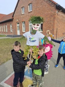Dwoje dzieci trzyma dużą kukłę ze słomy i gałązek, symbol zimy, na terenie szkolnym.