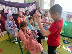 Uczeń wręcza koleżance upominek, a uczennice siedzą w tle podczas szkolnego świętowania Dnia Kobiet.