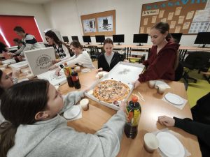 Uczniowie siedzą przy stole w klasie i dzielą się pizzą podczas szkolnych obchodów Dnia Kobiet.