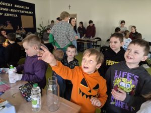 Grupa uczniów klas 0–3 zgromadzona przy stanowisku doświadczalnym. Chłopiec w pomarańczowej bluzie wskazuje ręką na doświadczenie. Na stole znajdują się butelka z wodą, szklany dzbanek i materiały do eksperymentów. W tle widoczni są uczniowie klas 7 i 8 oraz opiekunowie.