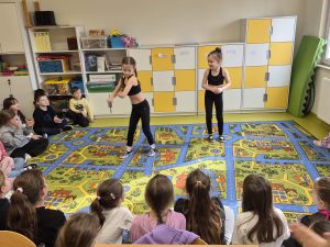 Uczniowie klas 1–3 siedzą w kręgu w sali lekcyjnej i oglądają występ taneczny dwóch dziewczynek prezentujących swoje hobby – taniec.