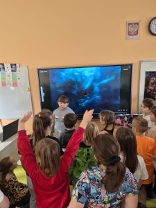 Uczniowie klas 1–3 stoją przed ekranem multimedialnym i oglądają prezentację wideo związaną z hobby jednego z uczniów.