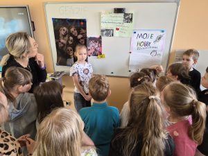 Uczennica klas 1–3 stoi przed tablicą i opowiada o swoim hobby, a pozostali uczniowie oraz nauczycielka słuchają prezentacji.