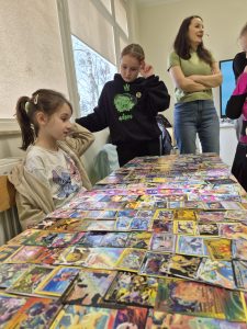 Uczniowie klas 1–3 oglądają rozłożoną na stole kolekcję kart kolekcjonerskich jako przykład hobby związanego z grami i zbieraniem kart.