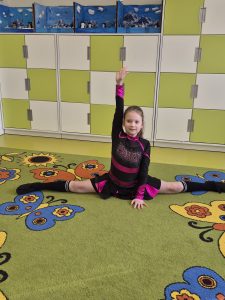 Uczennica klas 1–3 prezentuje swoje hobby sportowe, wykonując szpagat na dywanie w sali lekcyjnej.