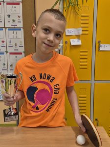 Uczeń klas 1–3 trzyma puchar i rakietkę do tenisa stołowego, prezentując hobby związane ze sportem.