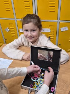 Uczennica klas 1–3 siedzi przy stoliku i prezentuje pudełko z koralikami i biżuterią jako hobby związane z rękodziełem.