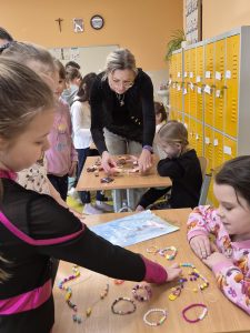 Nauczycielka i uczniowie klas 1–3 oglądają na stolikach kolorowe bransoletki wykonane z koralików podczas prezentacji hobby plastycznego.