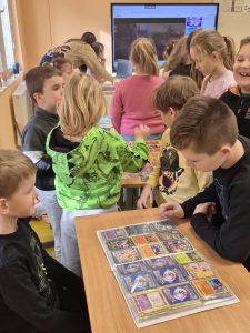 Uczniowie klas 1–3 stoją przy stolikach i oglądają albumy z kartami kolekcjonerskimi, rozmawiając o swoim hobby.