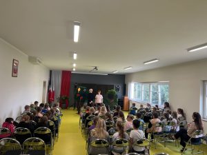 Uczniowie siedzą na krzesłach i oglądają przedstawienie teatralne na szkolnej scenie.